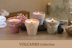 Four lit wax candles in rustic volcano-shaped holders on a wooden surface, with a spa-like bathroom setting in the background and the text 'VOLCANES Collection' at the bottom.