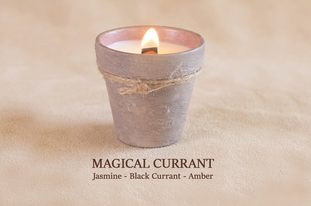 A small lit candle in a rustic gray ceramic pot with a twine band, set on a beige textured surface. Text below reads 'MAGICAL CURRANT Jasmine - Black Currant - Amber'.