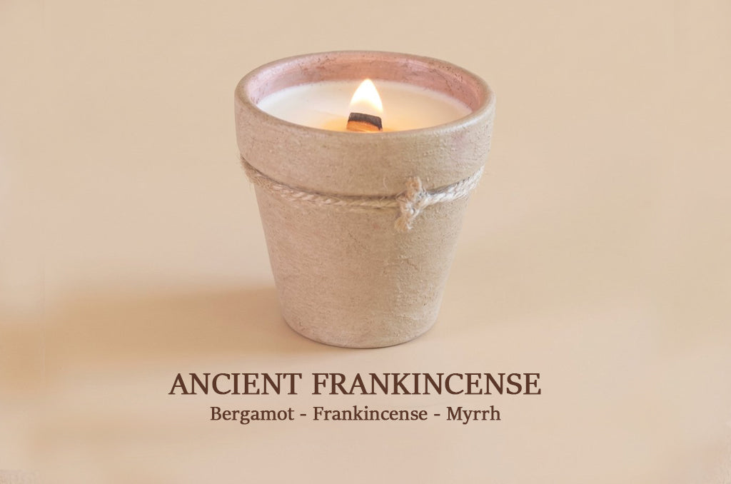 A lit candle in a small textured beige pot with a visible flame, centered on a warm beige background. Text below reads 'ANCIENT FRANKINCENSE' with 'Bergamot - Frankincense - Myrrh'.