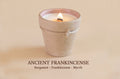 A lit candle in a small textured beige pot with a visible flame, centered on a warm beige background. Text below reads 'ANCIENT FRANKINCENSE' with 'Bergamot - Frankincense - Myrrh'.
