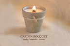 A small lit candle in a gray-green terracotta pot with twine, set on a beige fabric background. Text below reads 'GARDEN BOUQUET' with 'Peony - Magnolia - Freesia'.