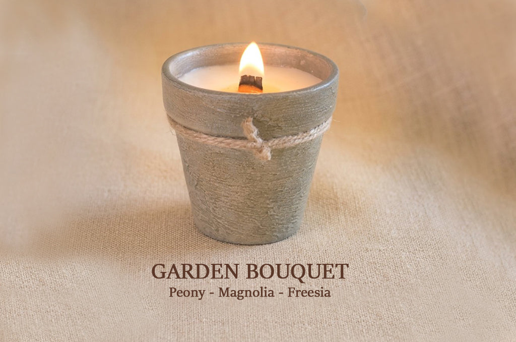 A small lit candle in a gray-green terracotta pot with twine, set on a beige fabric background. Text below reads 'GARDEN BOUQUET' with 'Peony - Magnolia - Freesia'.