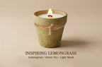 A single lit candle in a textured, terracotta-colored ceramic pot with a natural jute twine bow around the middle, set against a solid beige background. Text below reads 'INSPIRING LEMONGRASS' and 'Lemongrass - Green Tea - Light Musk'.
