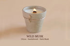 A lit candle in a textured, light gray ceramic pot with a small twine bow, set against a warm beige background. Text below reads 'WILD MUSK Citrus - Sandalwood - Dark Musk'.