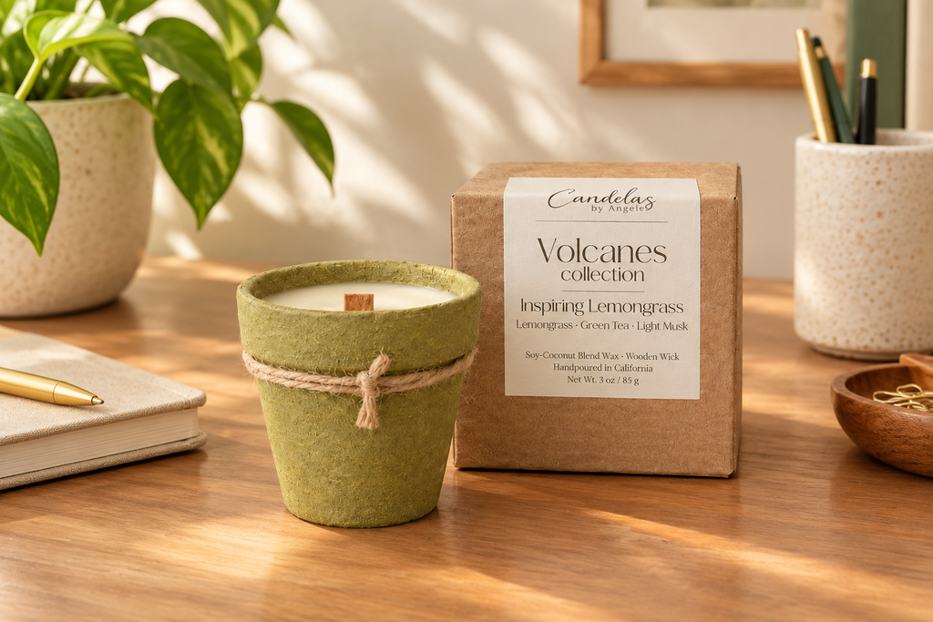 A candle in a textured sage green pot with a twine band sits on a wooden desk beside its kraft paper box labeled 'Volcanes collection'. The scene is styled with a potted plant, a notebook, and stationery in a warm, sunlit room.