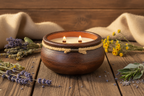 A rustic brown ceramic candle holder with two lit candles, decorated with a rope band and a small metal accent, set on a wooden table with dried lavender, sage, and yellow flowers.
