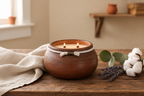 A rustic brown ceramic candle holder with two lit candles sits on a wooden table, styled with a beige linen cloth, lavender sprigs, and cotton bolls in a warm, softly lit room.